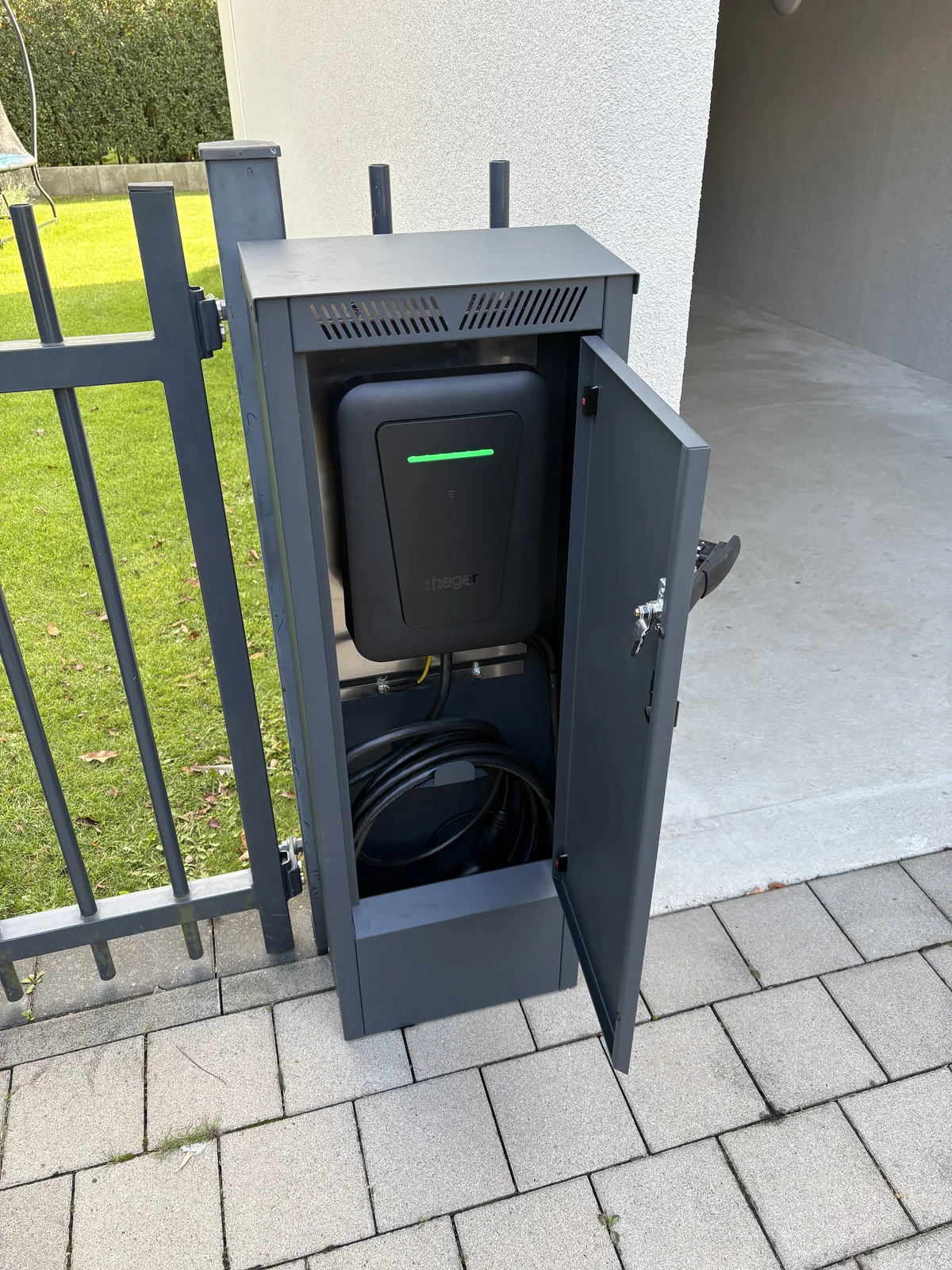 Wallbox mit grüner Statusanzeige und Ladekabel in einem geöffneten grauen Metallgehäuse neben einer Einfahrt.