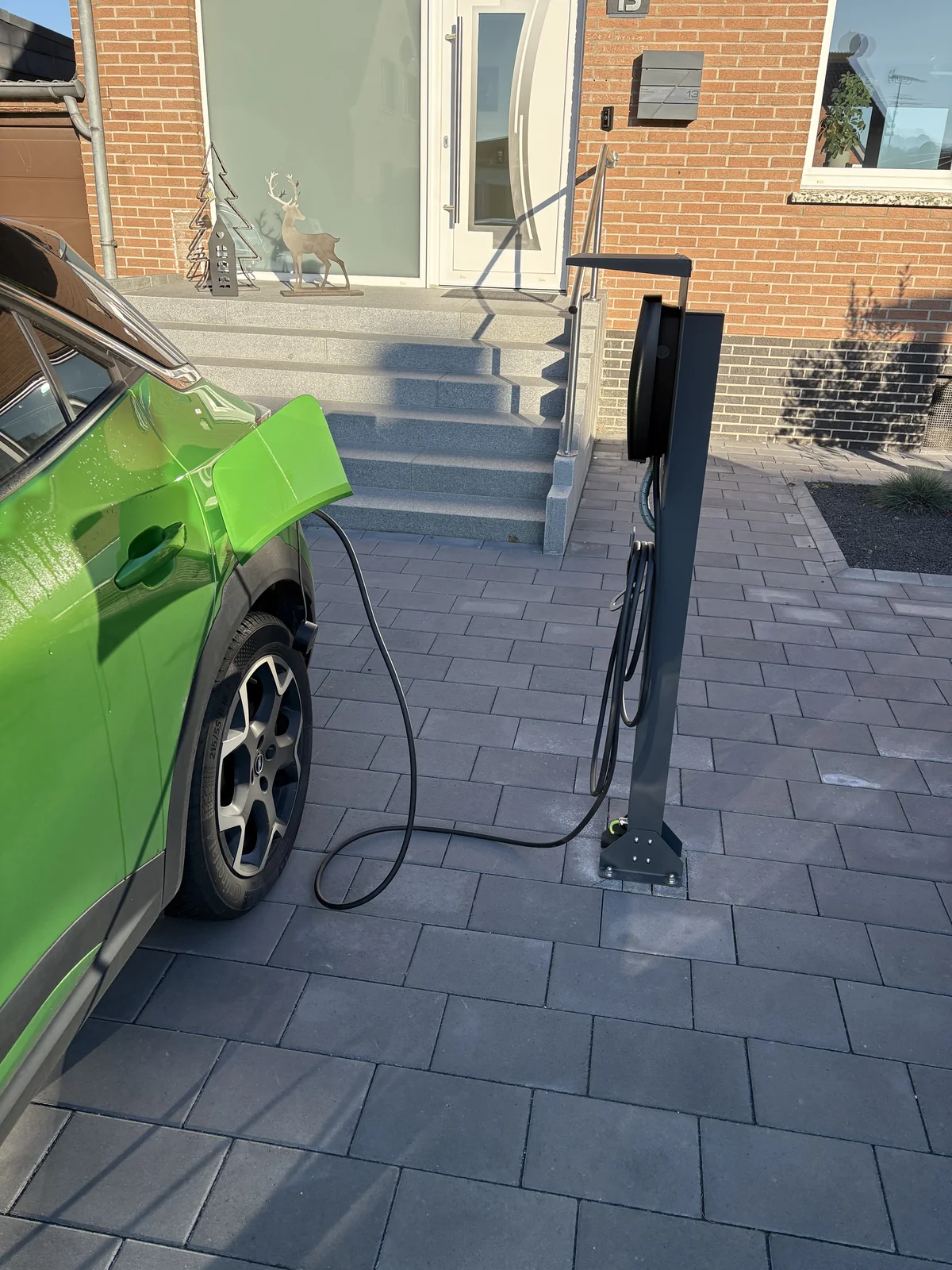 Freistehende Wallbox auf einer Standsäule neben einem grünen Elektroauto am gepflasterten Stellplatz.